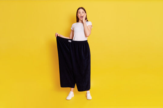 Caucasian shocked female with dark hair looking at camera with big eyes and open mouth, covering cheek with palm, wearing too big pants, woman lost weight, isolated over yellow background.