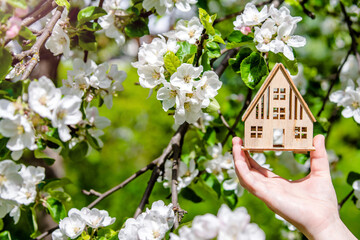 The girl holds the house symbol against the background of blossoming appletree
