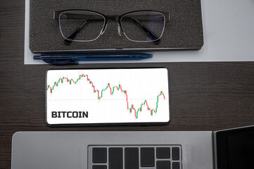 Buy or sell Bitcoin concept. Top view of stocks price candlestick chart in phone on table near laptop, notepad and glasses with inscription Bitcoin. Business, finance concept.