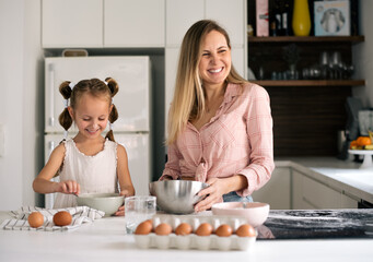 Mother and daughter together in kitchen prepare dough for favorite cake. On table is mixer and ingredients for cookies: flour, eggs, sugar, milk. Fun to cook with mom. Grandma's recipe.