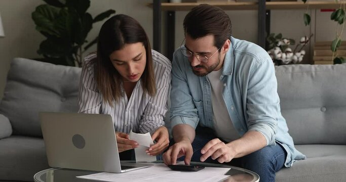 Young married couple sit on couch at home calculating expenses using calculator feel upset and stressed discussing money overspend. Lack of finances, high taxes, debt and crisis, bankruptcy concept