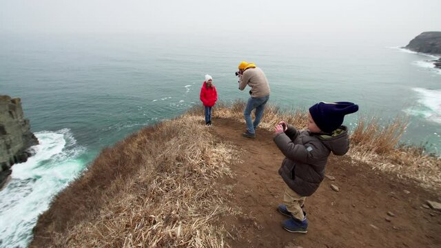 4 years old boy is taking photo or video of marine landscape. Father is taking photos of his daughter who is standing on the cliff edge