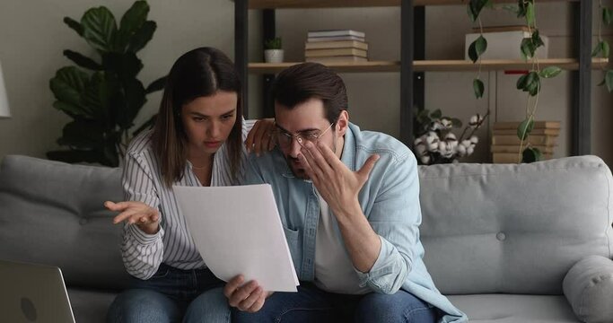 Young stressed married couple sit on couch hold papers reading bad news in legal document, got warning notice feel shocked looking frustrated. Pay penalty, unpaid debt, eviction notification concept