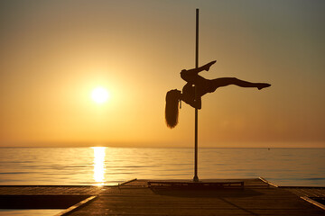 pole dance and acrobatic pole poses in chiaroscuro. Pier, Baltic Sea, sunrise and orange sky.
