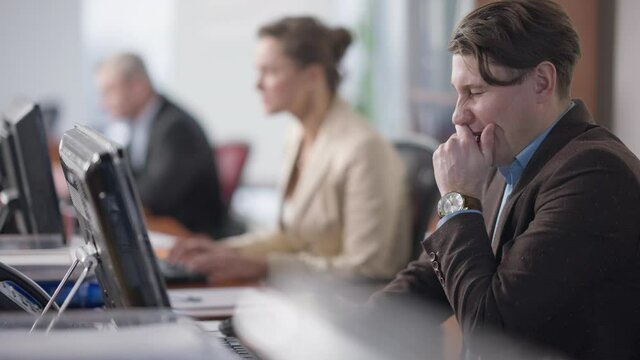 Tired handsome overworked man yawning typing on computer keyboard with busy blurred coworkers working online at background. Exhausted Caucasian employee at workplace in office