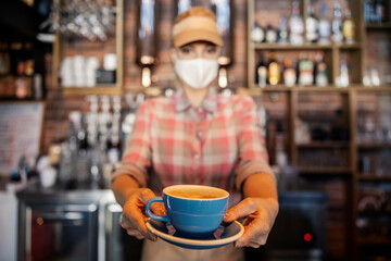 Coffee during the corona. A portrait of a woman with a mask standing in a bar provides a cup of coffee in front of the camera, focusing on a cup of coffee. Coffee shop, corona virus