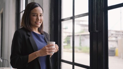 Asian business woman in a good mood smile thinking with holding coffee cup, man standing near window looking at camera. Successful young business people concept.