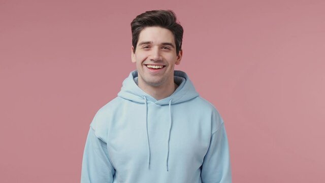 Handsome stylish brunet young man 20s wears blue streetwear hoodie posing looking camera asides charming smile isolated on pastel pink wall background studio portrait People emotions lifestyle concept