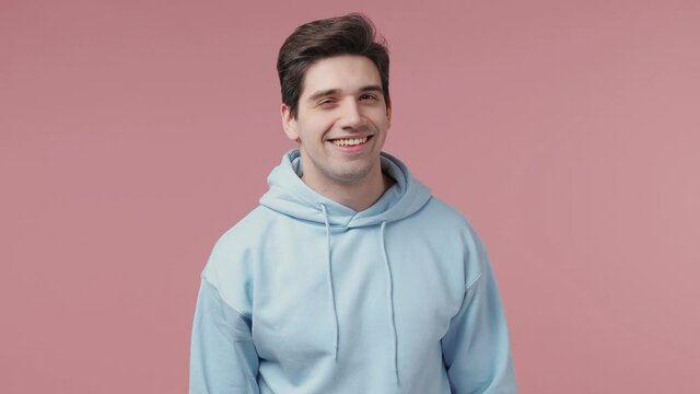 Handsome brunet young man 20s wears blue streetwear hoodie looking camera asides charming smile wink eye blink isolated on pastel pink wall background studio portrait People emotions lifestyle concept