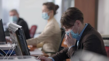Side view of ill Caucasian man in coronavirus face mask sneezing and blurred coworkers leaving at background. Frightened office workers walking away from sick colleague. Scare on Covid-19 pandemic