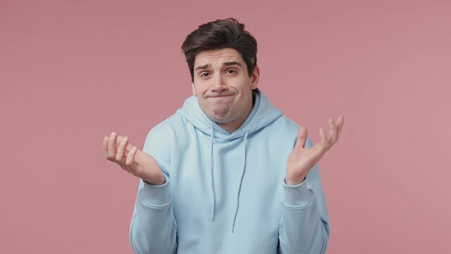 Fun confused shy shamed young man 20s wear blue streetwear hoodie look camera spreading hands say oops oh i am so sorry isolated on pastel pink wall background studio portrait People lifestyle concept