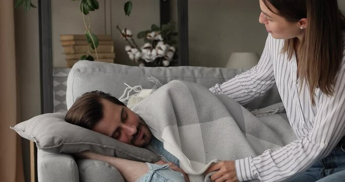 Tired overworked husband falling asleep on couch in living room loving wife cover sleeping beloved man with warm plaid expressing care, caring about healthy and comfort rest. Cherish and love concept