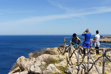 Group of hikers observing Punta Nati Lighthouse from the Sa Falconera point of view, in Menorca (Balearic Islands, Spain)