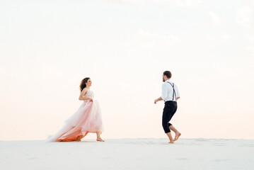 Fototapeta premium young couple a guy in black breeches and a girl in a pink dress are walking along the white sand