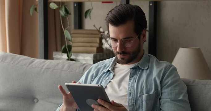 Busy attractive man in glasses sit on sofa holding tablet device. Do telecommute job remotely, chatting in social media, choose goods on web stores. E-commerce, virtual services, fun app user concept