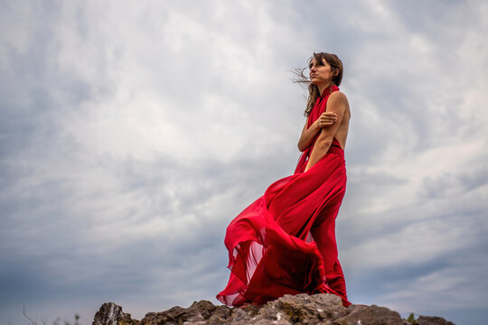 A woman in a red dress stands above a stormy sky, her dress fluttering, the fabric flying in the wind.