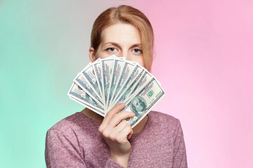 Portrait of beautiful caucasian woman holding dollars banknotes in hand and covering her face with bunch of money bills over green and red background.