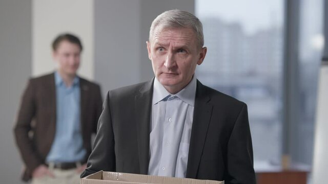 Portrait of fired stressed senior man holding box standing in office. Upset Caucasian grey-haired employee indoors. Age discrimination in business and frustration concept