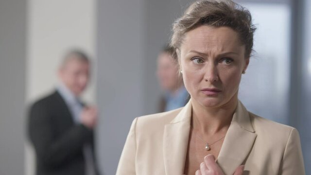 Stressed brunette woman with hazel eyes standing indoors thinking with blurred men talking at background. Portrait of bullied Caucasian female employee in office. Discrimination and harassment