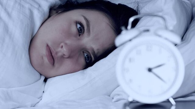Insomnia, anxiety, stress. Face of sleepy woman at night, she looking tiredly at alarm clock standing next to it, focus of camera switches from clock to face of woman. Close-up