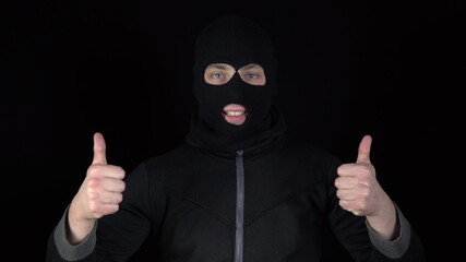 A man in a balaclava mask shows his approval mark with his hands. The thug raised his hands and showed his thumbs up against a black background.