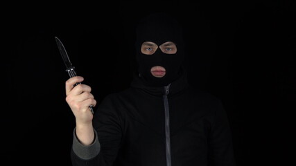 A man in a balaclava mask stands with a butterfly knife. The thug twists a knife on a black background.