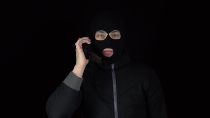 A man in a balaclava mask with a phone from the 90s. The bandit is talking on the phone. On a black...
