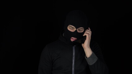 A man in a balaclava mask is talking on a phone from the 90s. A thug screaming on the phone on a black background.