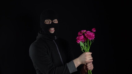 A man in a balaclava mask is standing with flowers. Bandit sniffs a bouquet of pink flowers on a...