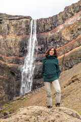 Naklejka na ściany i meble Hengifoss, Iceland »; August 2017: A young woman with a green sweater under the Hengifoss waterfall sitting on a stone.