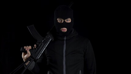 A man in a balaclava mask stands with an AK-47 assault rifle. A bandit stands with a gun on his shoulder on a black background.