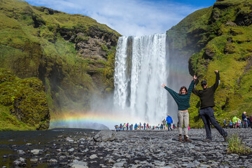 A couple at the Skogafoss waterfall on a summer afternoon with a rainbow and tourists under the...