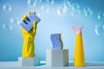 Creative still life with supplies for cleaning or housekeeping on podiums over blue background.Fenmale hand in yellow rubber glove with clean sponge, spray bottle and color sponges..