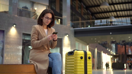 A young woman sits with a phone in her hands in the airport with a suitcase.