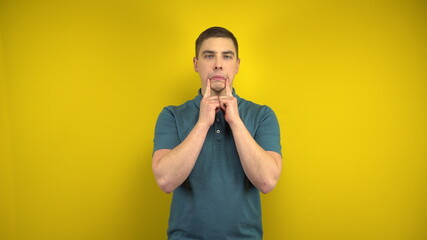 A young man makes a sad smile on her face with his fingers on a yellow background. Man in a green...
