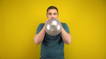 A young man inflates a silver balloon with his mouth on a yellow background. Man in a green polo.