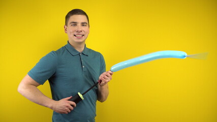 A young man inflates a long blue balloon with a pump on a yellow background. Man in a green polo.