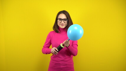 Fototapeta na wymiar A young woman inflates a blue balloon with a pump on a yellow background. Girl in a pink turtleneck and glasses.