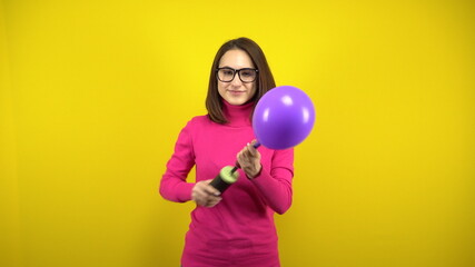 Fototapeta premium A young woman inflates a purple balloon with a pump on a yellow background. Girl in a pink turtleneck and glasses.