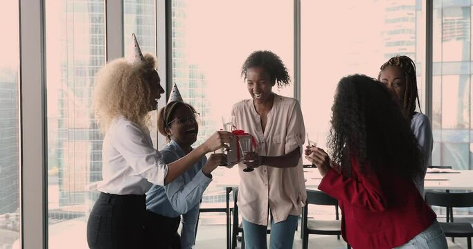 Five women gather in office, colleagues congratulate African mate gives gift box clinking glasses with champagne laughing enjoy event at workplace. Friendly work relation, birthday celebration concept