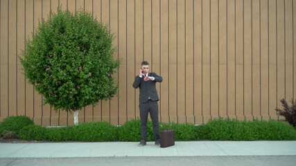 A young businessman in a suit speaks on the phone. Serious man standing with briefcase in hand on background of the building with a flowerbed.