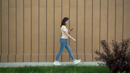 A young woman goes with a phone in her hand. A girl walks near the building and carries on correspondence in a smartphone.