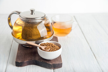 uncooked buckwheat in white plate and wooden scoop, on a white background close-up. Buckwheat tea and granules. Tea in transparent teapot. Space for text.
