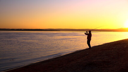 A man is fishing at sunrise by the river. The yellow sun rises from the horizon. Silhouette of a...