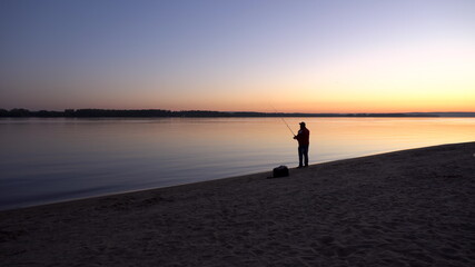 Fototapeta premium A man is fishing at sunrise by the river. The yellow sun rises from the horizon. Silhouette of a fisherman.