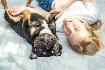 girl lies on the bed in an embrace with her dog of the French Bulldog breed