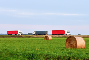 View of a field with hay in rolls against the background of trucks with semi-trailer driving along the highway. Harvesting dry grass for agriculture. Lorry on the road