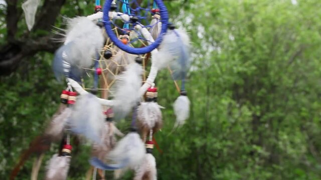 A dreamcatcher with multi-colored feathers flutters in the wind against the backdrop of blurry greenery of nature. Relax concept.