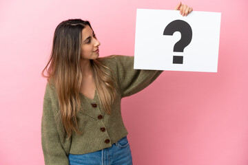 Young caucasian woman isolated on pink background holding a placard with question mark symbol