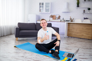 Handsome young man sitting on the floor and holding bottle of water while having online workout